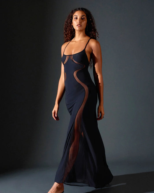 A woman modeling a floor-length navy gown with sheer mesh side panels and thin straps, photographed against a dark gray studio background.